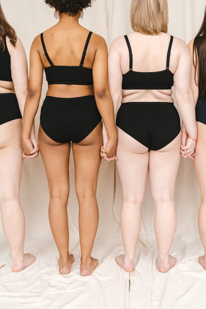 A view behind several women of various body types in comfortable black underwear, holding hands.